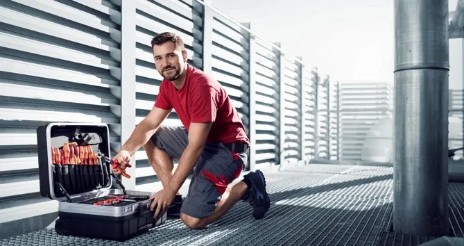 Ein Techniker in rotem T-Shirt und grauer Arbeitskleidung kniet auf einem Gitterboden in einer modernen technischen Anlage. Vor ihm steht ein geöffneter Werkzeugkoffer, im Hintergrund sind Lüftungsschächte und Rohre zu sehen.